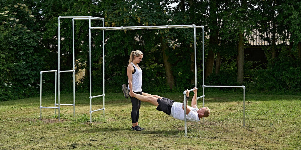 Udendørs træning | Mand laver Rows på havestativ med hjælp fra kvindelig træningspartner